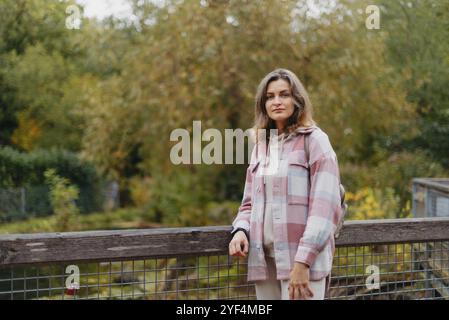 Ritratto di carina giovane donna in abbigliamento casual in autunno, in piedi sul ponte sullo sfondo di un parco autunnale e fiume. Bella donna che cammina a Par Foto Stock