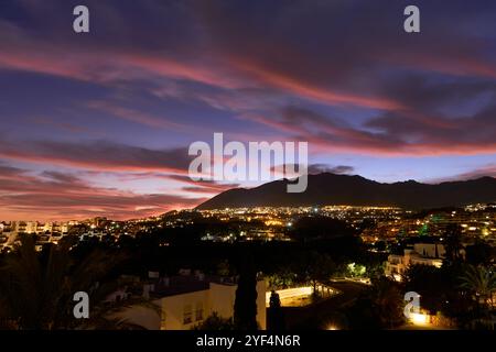 Splendido tramonto dietro una montagna in una zona residenziale con vegetazione lussureggiante, nuvole rosse e crepuscolo con le luci degli edifici che si accendono Foto Stock