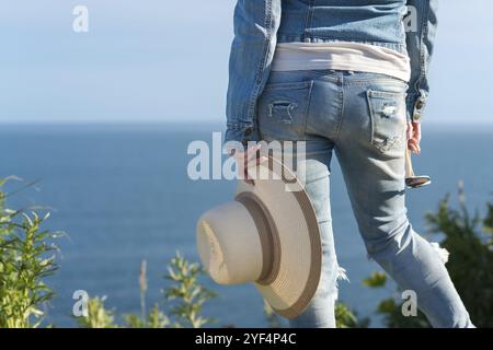 Vista posteriore tagliata sotto la vita sulle gambe della donna in jeans blu. Ragazza hipster irriconoscibile che tiene cappello di paglia in una mano e occhiali da sole in un'altra, sta Foto Stock