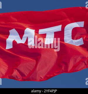 Bandiera rossa che sventola nel vento con testo in russo: MTS Mobile TeleSystems, abbreviazione operatore di telefonia mobile Federazione Russa. Bandiera del marchio su sfondo blu S Foto Stock