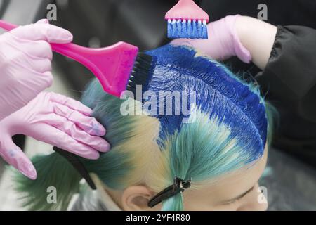 Due parrucchieri che utilizzano pennello rosa mentre si applica vernice blu alla cliente femminile con colore smeraldo dei capelli, durante il processo di tintura dei capelli in lapide unico la Foto Stock