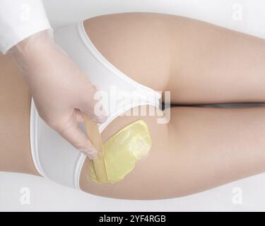 Vista dall'alto della mano nel guanto applicazione di cera calda verde sulle natiche di donna snella utilizzando la spatola. Processo di depilazione con cera calda in salone di bellezza professionale Foto Stock