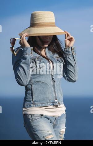 Ritratto di una donna hipster con giacca in denim e jeans che si nascondono con un cappello di paglia sul viso. Donna adulta che posa sullo sfondo di un cielo blu senza nuvole e Foto Stock