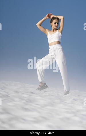 Donna adulta alla moda che posa le mani in alto, gli occhi chiusi, in piedi attraverso la neve con la nebbia leggera nelle giornate di sole con il cielo blu. Modello di moda biondo vestito Foto Stock