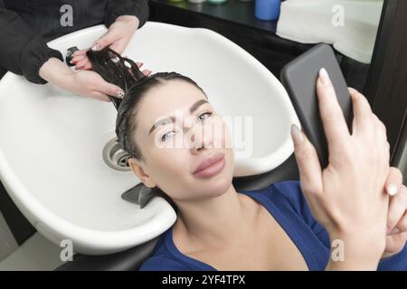 La bruna guarda lo smartphone mentre il parrucchiere è occupato a lavarsi i capelli al salone di bellezza. Il parrucchiere lava i capelli lunghi del cliente con Sha Foto Stock