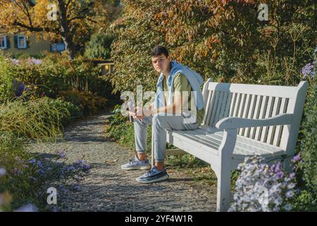 Un adolescente siede su una panchina nel parco beve il caffè da una tazza termica e guarda un telefono. Ritratto di un bel ragazzo allegro seduto sulla panchina libera Foto Stock