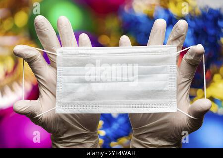 Mani del medico che tengono la maschera in guanti medici bianchi su sfondo bokeh palle di Natale decorazioni. Concetto di protezione per felice anno nuovo e. Foto Stock