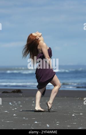 Donna rossa con lungo abito marrone viola in piedi su una spiaggia sabbiosa con la testa rivolta verso l'alto e gli occhi chiusi, prendendo il sole sullo sfondo del mare blu in estate Foto Stock