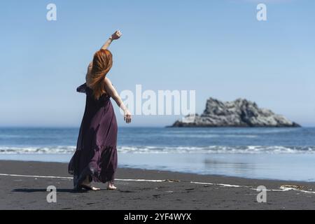 Irriconoscibile donna rossa in abito scuro e puce lungo in piedi sulla spiaggia sabbiosa con una mano sollevata sullo sfondo del mare blu, isola vicino alla riva sul summ Foto Stock