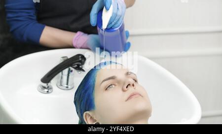 La parrucchiere mette le mani nei guanti spreme lo shampoo dal tubo alla testa giovane donna con i capelli blu mentre lava i capelli in lavello speciale e doccia presso Beauty sa Foto Stock