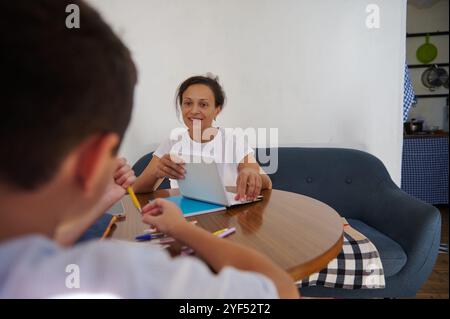 Una madre aiuta suo figlio con i compiti a un tavolo di legno, creando un ambiente di apprendimento accogliente. La scena cattura il legame e il supporto educativo con Foto Stock