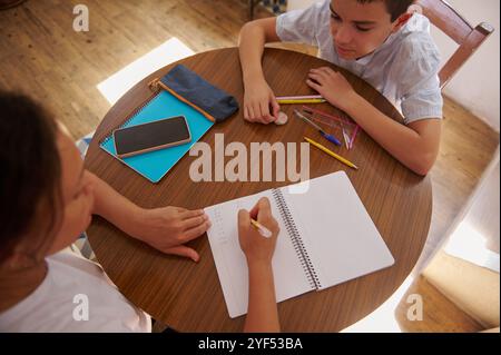 Una madre aiuta suo figlio con i compiti, scrivendo su un quaderno su un tavolo di legno. Sono circondati da forniture scolastiche e si concentrano sul compito, creato Foto Stock