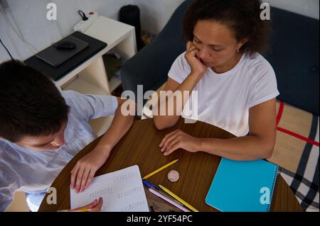 Una madre siede a un tavolo con suo figlio, aiutandolo con i compiti di matematica. Il ragazzo si sta concentrando sul foglio di lavoro, circondato da matite e quaderno Foto Stock