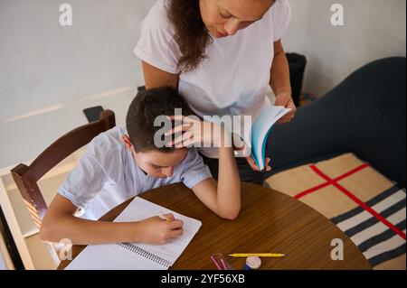 Una madre aiuta suo figlio con i compiti a un tavolo di legno rotondo. Il bambino si concentra sulla scrittura, mentre la madre fornisce una guida, favorendo una suppo Foto Stock