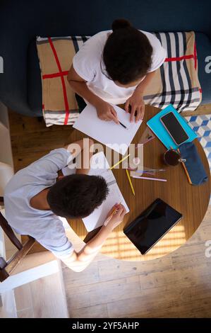 Due studenti che lavorano insieme a casa su un progetto. Vista dall'alto di un tavolo con documenti, tablet e cancelleria, che mostra il lavoro di squadra e gli studenti Foto Stock