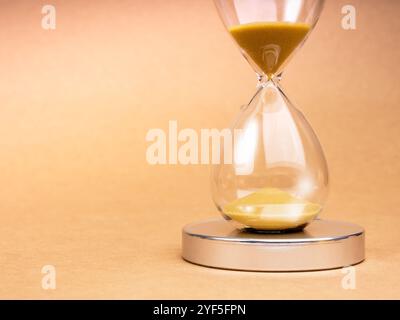 Clessidra trasparente da primo piano con sabbia dorata su podio cromato su sfondo marrone chiaro riciclato con spazio per le copie. Sabbia dorata in vetro di sabbia isolata o Foto Stock