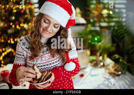 Natale. Felice elegante donna d'affari di 40 anni con cappello di babbo Natale e maglione rosso con biscotti tradizionali che parla su uno smartphone Foto Stock