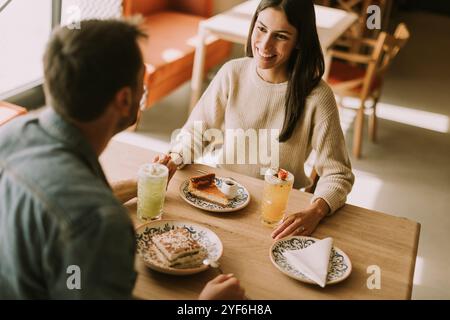 In un caffè caldo e invitante, una coppia condivide risate e deliziose prelibatezze, assaporando caffè e pasticcini mentre si crogiola al sole del pomeriggio. Foto Stock