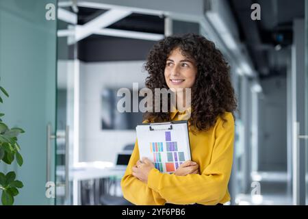 Donna d'affari sicura con capelli ricci in un ufficio moderno contiene appunti con grafici colorati. Irradia ispirazione e attenzione in un ambiente professionale. Foto Stock
