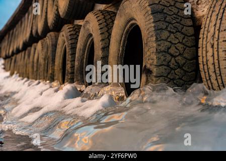 Grandi blocchi di ghiaccio ghiacciati su un porticciolo con pneumatici per auto in inverno. Il tramonto dorato si illumina sul lato di un lago ghiacciato in inverno. Foto Stock