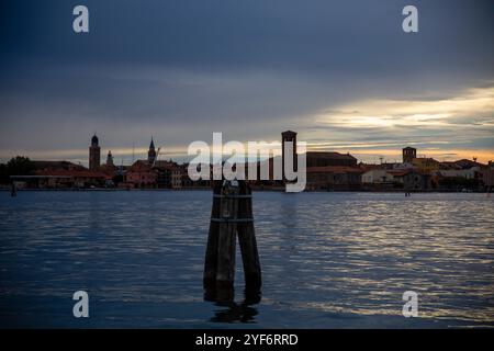 Una bricola veneziana in legno (limite del canale navigabile) nella laguna di Chioggia, nell'azzurro del tramonto, con le case sullo sfondo, Chiogg Foto Stock