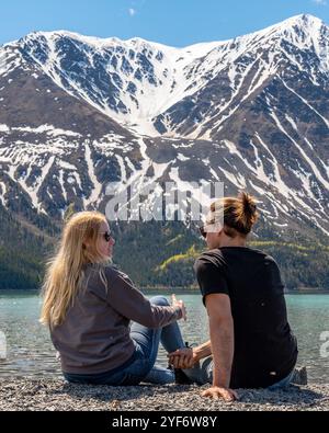 Due persone, amici, coppia seduti su sedie da campeggio con tavolo da picnic sull'area panoramica della riva del lago nel territorio dello Yukon, Canada settentrionale, in primavera Foto Stock