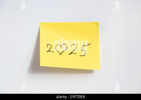 2025 dove al posto dello zero un cuore è disegnato a mano su un pezzo di carta giallo su sfondo bianco, felice anno nuovo 2025 Foto Stock