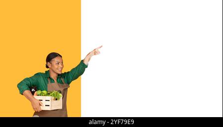Donna sorridente con un cesto di verdure punti con entusiasmo al chiuso Foto Stock