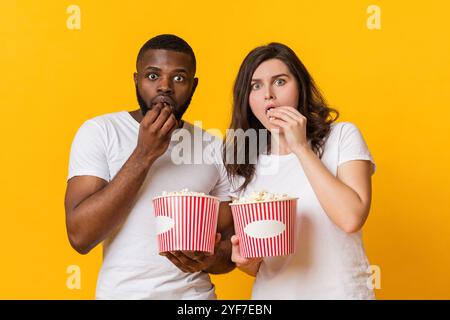 Ritratto di coppia interracial dazed che mangia popcorn con espressione di faccia spaventata Foto Stock