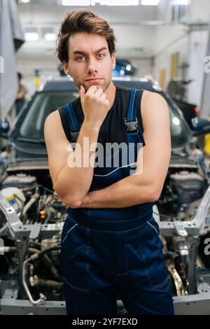 Ritratto verticale di un meccanico premuroso maschio in uniforme che guarda la fotocamera non sa quale riparazione durante la manutenzione dell'auto in officina. Foto Stock