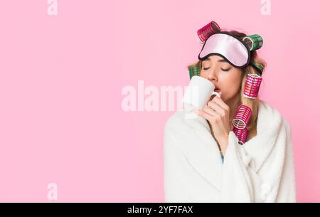 Donna assonnata in arricciacapelli e maschera per il sonno avvolto in coperta e bevande caffè da tazza Foto Stock