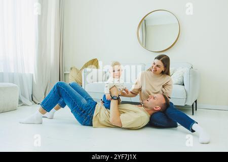 Una giovane coppia di sposi si diverte a giocare con un bambino piccolo in un moderno soggiorno insieme. Genitori sorridenti mamma e papà si divertono a trascorrere il tempo Foto Stock