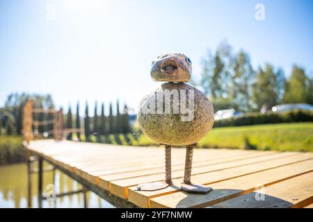 Una scultura di uccello in metallo sorge su un molo di legno accanto a uno stagno, con alberi e un cielo blu sullo sfondo, suggerendo una giornata di sole in un parco o in un giardino. Foto Stock