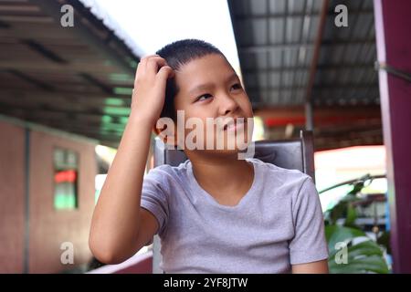 Un giovane ragazzo premuroso siede all'aperto con una camicia grigia casual, che riflette sulla vita. La sua espressione trasmette un senso di innocenza e curiosità. Perfetto per Foto Stock