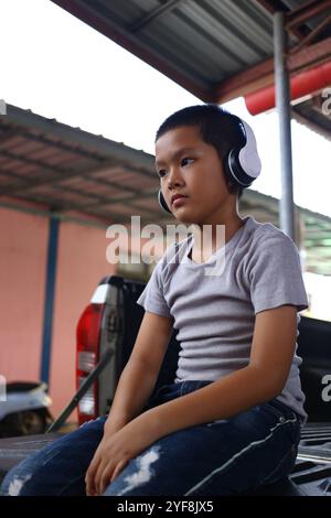 Un ragazzo premuroso che indossa le cuffie si siede casualmente all'aperto, perdendo nella sua musica. Il momento cattura l'innocenza e la tranquillità dell'infanzia in un set semplice Foto Stock