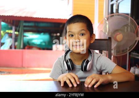 Un ragazzo che indossa le cuffie sorride brillantemente mentre è seduto al chiuso. L'atmosfera e' vivace, riflettendo uno spirito giocoso e giovanile, ideale per fami Foto Stock