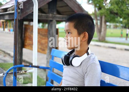 Un ragazzo siede da solo su una panchina blu e indossa le cuffie, ascoltando musica in un tranquillo parco all'aperto, circondato dalla natura e dalla luce del sole. Foto Stock