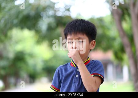 Un ragazzo si trova in un ambiente tranquillo all'aperto, sembrando preoccupato mentre tosse. La vegetazione lussureggiante che lo circonda accentua il suo momento di angoscia. Foto Stock