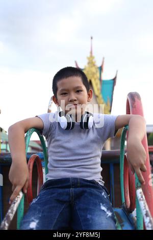 Un ragazzo allegro siede sulle attrezzature del parco giochi, indossa le cuffie e goditi un momento di divertimento e relax all'aperto, catturando l'essenza dei bambini Foto Stock