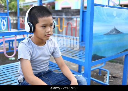 Un ragazzino siede all'aperto indossando le cuffie, perso nella sua musica. Il tranquillo parco giochi aggiunge un tocco di innocenza e tranquillità alla scena. Foto Stock