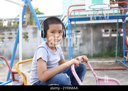 Un bambino siede sulle attrezzature del parco giochi, indossa le cuffie e ascolta la musica. La sua espressione riflette la calma e la soddisfazione in un ambiente all'aperto Foto Stock