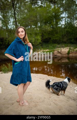 Giovane donna in Blue Poncho che si rilassa vicino al fiume Forest, Curious Dog che esplora le acque calme Foto Stock