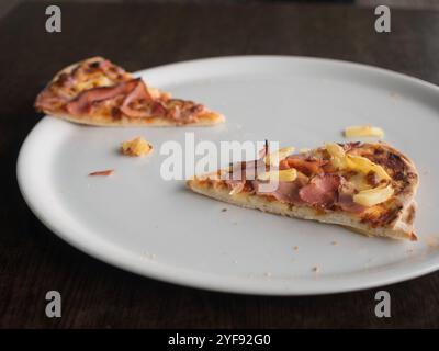 Due fette di prosciutto e pizza all'ananas su un grande piatto bianco sul tavolo con luce naturale. Foto Stock