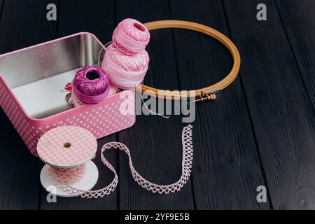 Filati assortiti e utensili da cucire in una scatola di latta rosa su sfondo nero in legno creano una disposizione accattivante per gli amanti dell'artigianato. Foto Stock