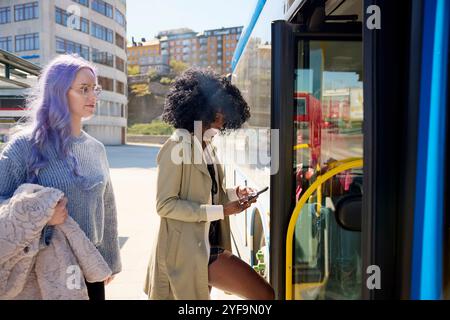 Donna felice dai capelli ricci che usa lo smartphone e salta in autobus con un'amica in strada Foto Stock