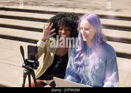 Felice donna di capelli viola che vatta con un'amica tramite smartphone mentre è seduta sui gradini della strada Foto Stock