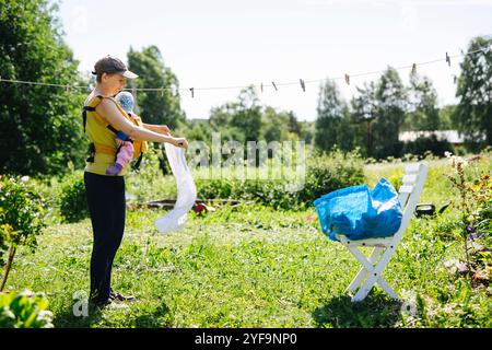 Vista laterale della madre che trasporta il bambino mentre appende il bucato sulla clothesline Foto Stock