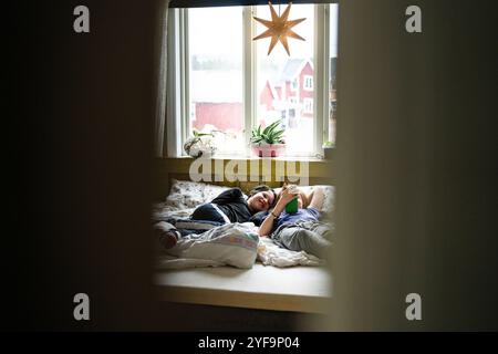 Un ragazzo sorridente sdraiato accanto a un fratello non binario che utilizza lo smartphone mentre è sdraiato a letto a casa Foto Stock