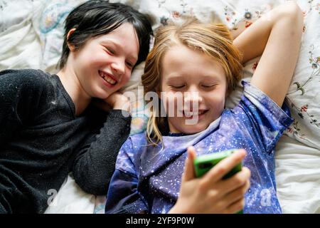 Fratello felice e fratello non binario che utilizza lo smartphone mentre si sdraia a letto a casa Foto Stock