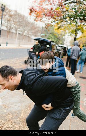 Papà dà a figlio un giro in piggyback in un'area urbana con foglie autunnali Foto Stock